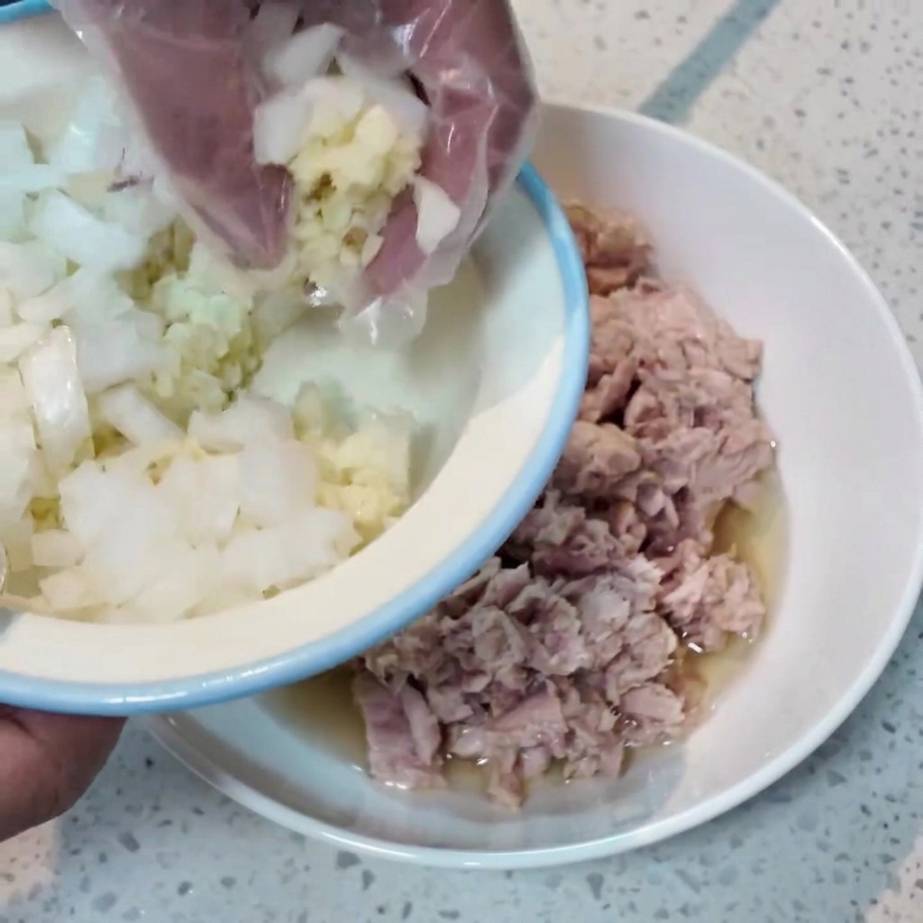A hand wearing a plastic glove transferring chopped onions and minced garlic from a small bowl into a larger bowl containing tuna.