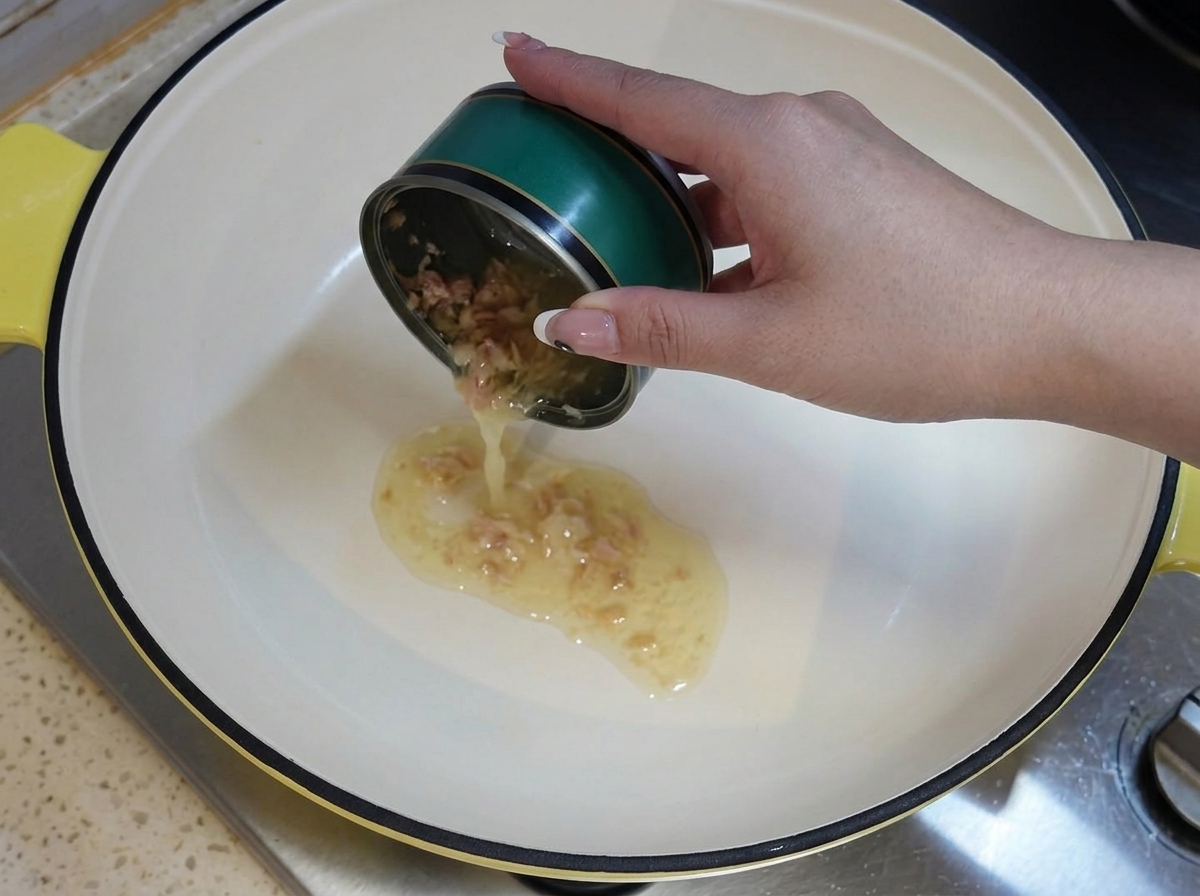 A hand holding an empty green tuna can, pouring its residual oil into a heated white pot on the stove.