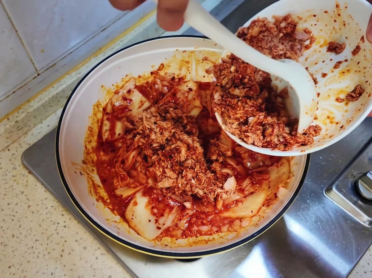Canned tuna mixture being scraped from a white bowl into the pan with kimchi using a white spoon.