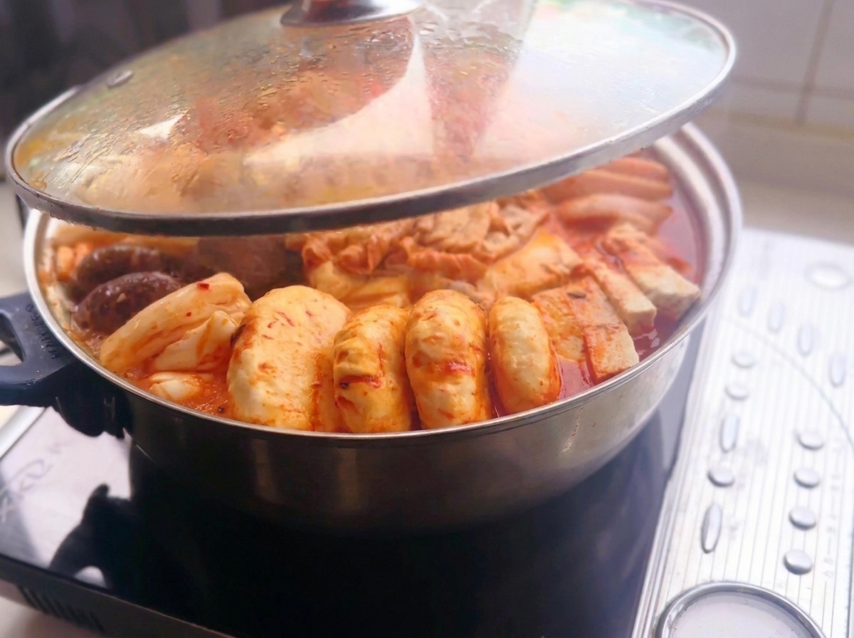 A glass lid being lifted off a metal hot pot filled with arranged ingredients like sliced meat, tofu, mushrooms, rice cakes, and dumplings in a spicy red broth on an induction cooker.