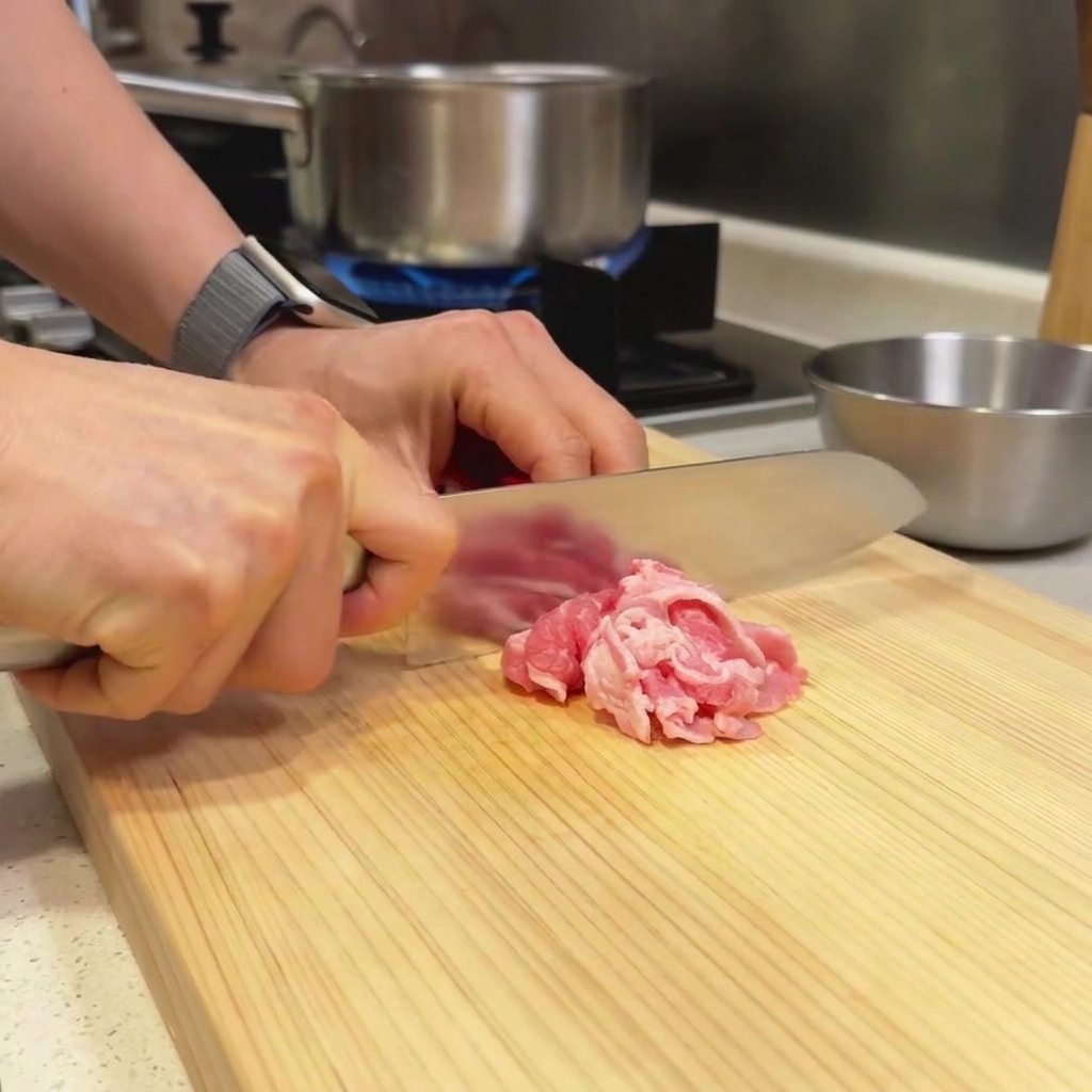 Thin slices of beef resting on a wooden cutting board being prepared with a chefs knife.