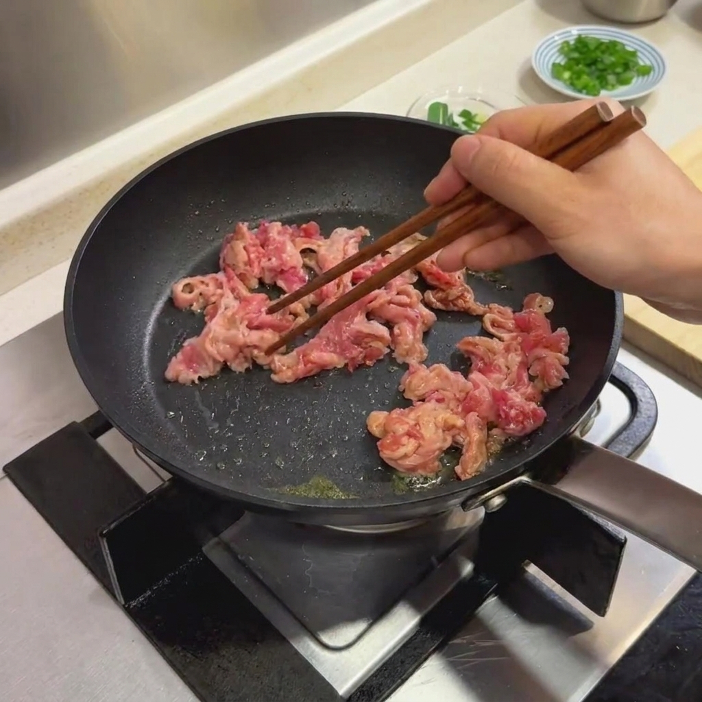 Marinated beef slices being cooked in a black frying pan using wooden chopsticks.