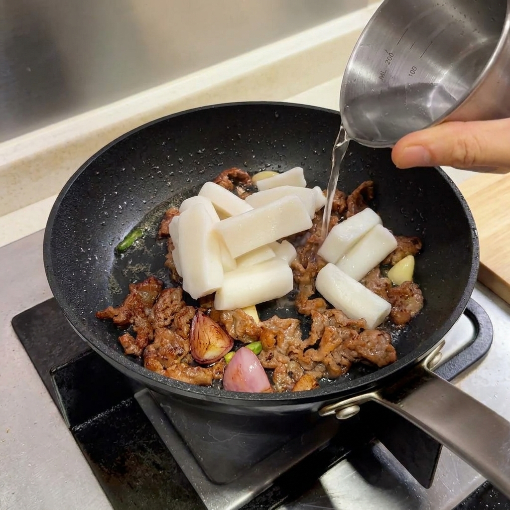 Rice cake cylinders in a frying pan with beef, being splashed with water from a small pitcher.