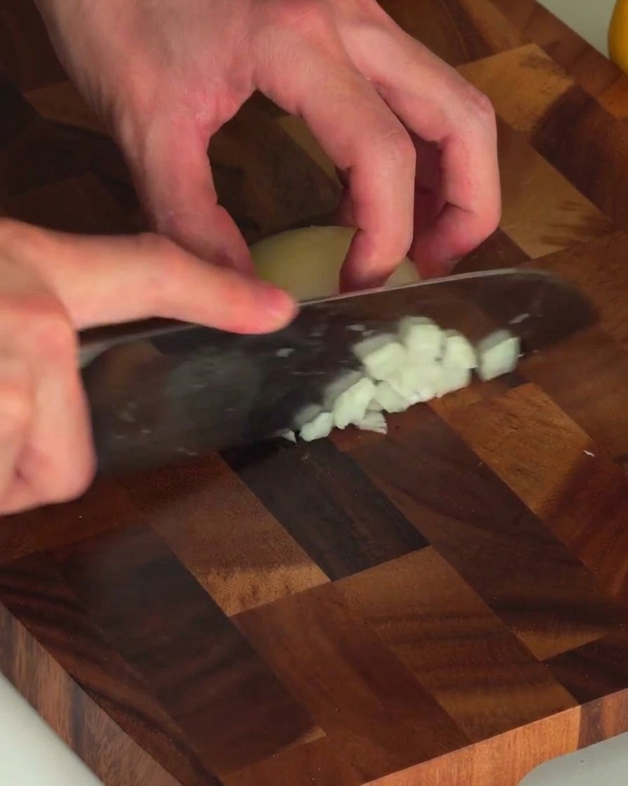 Hands holding a knife and finely chopping a white onion on a wooden board.