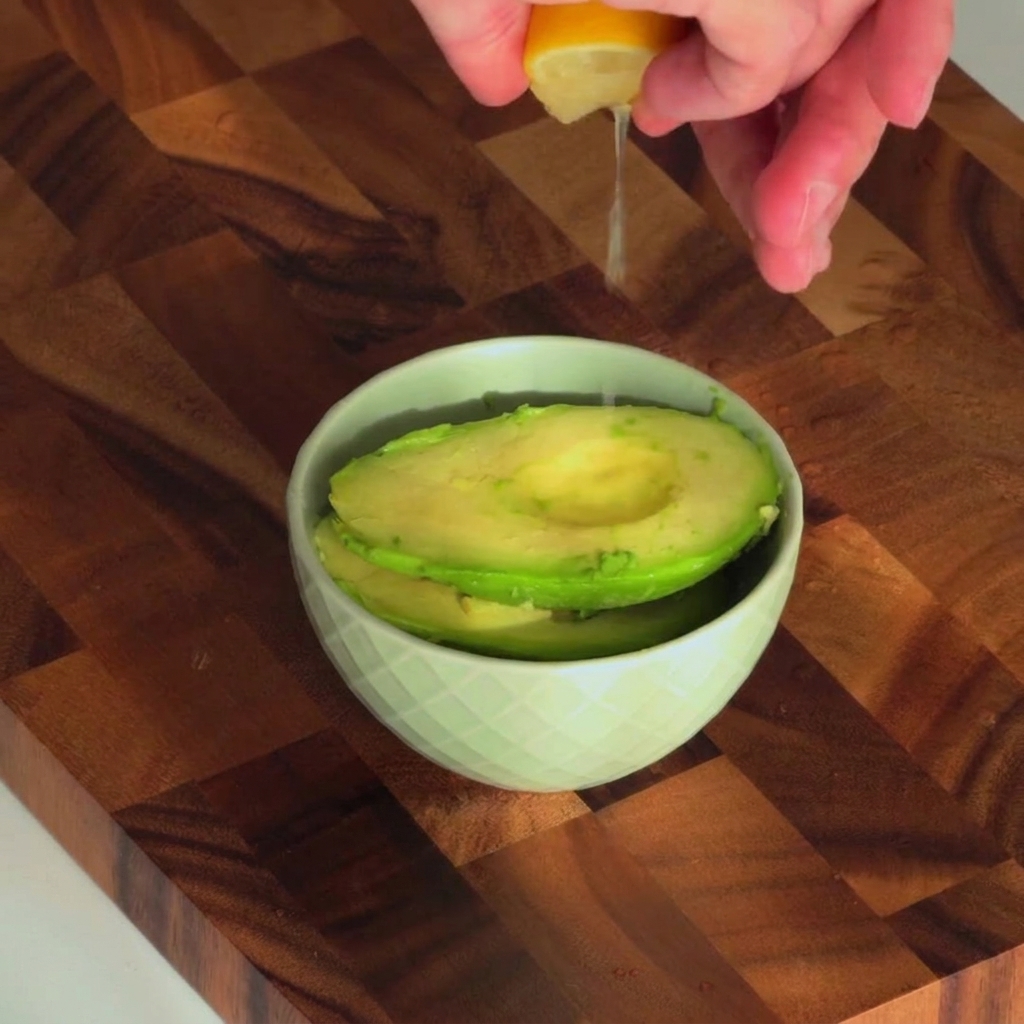 Fresh lemon juice being squeezed into a small bowl containing scooped avocado.