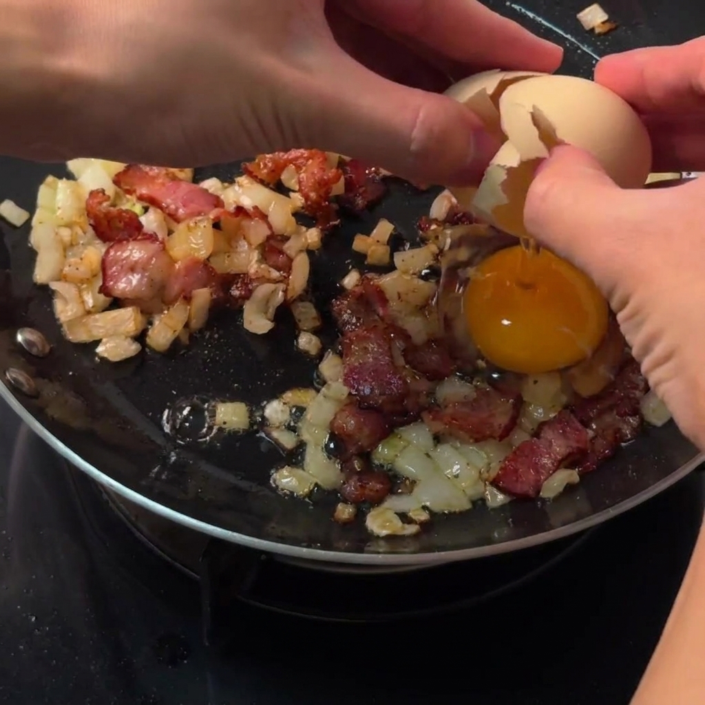 Hands cracking a raw egg directly into a cleared circular space among bacon and onions in a skillet.