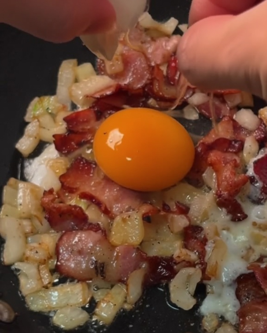 Fresh eggs being cracked into individual bacon and onion rings cooking in a skillet.