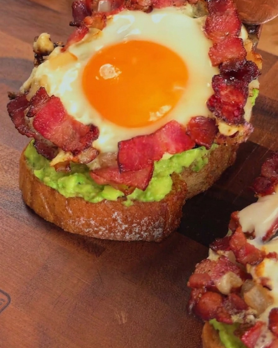 A cooked bacon and egg ring being layered over avocado toast with a wooden spatula.