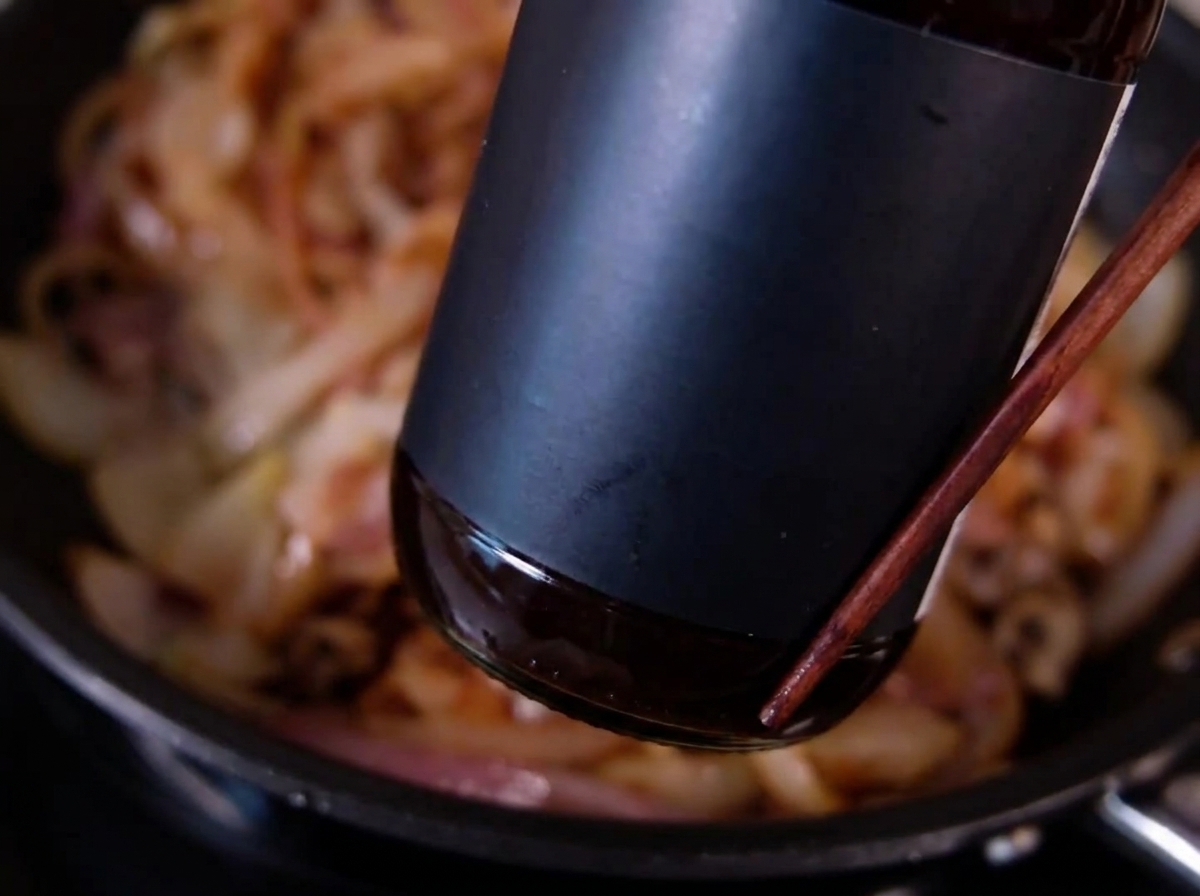 A dark bottle pouring sauce over thinly sliced, browned onions cooking in a pan.