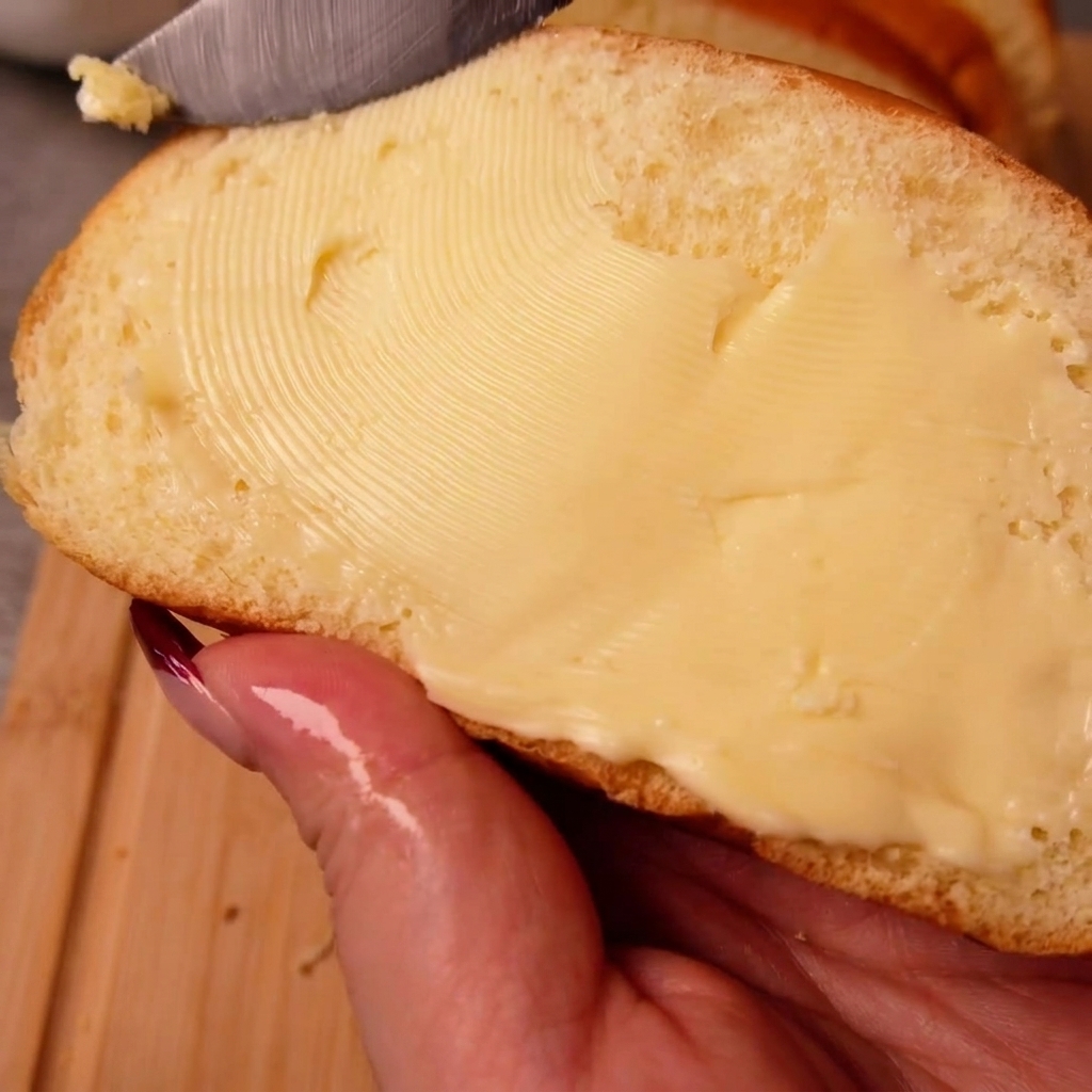 A hand holding a slice of white bread while spreading butter evenly across its surface with a knife.