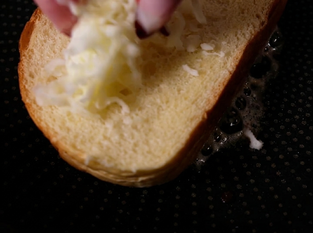 Fingers dropping shredded mozzarella cheese onto a plain slice of bread sitting in a hot pan.
