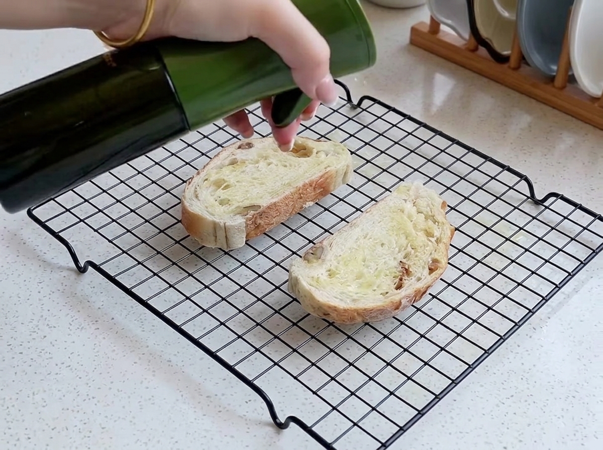 A hand spraying a fine layer of oil onto two slices of rustic bread laid out on a wire mesh rack.