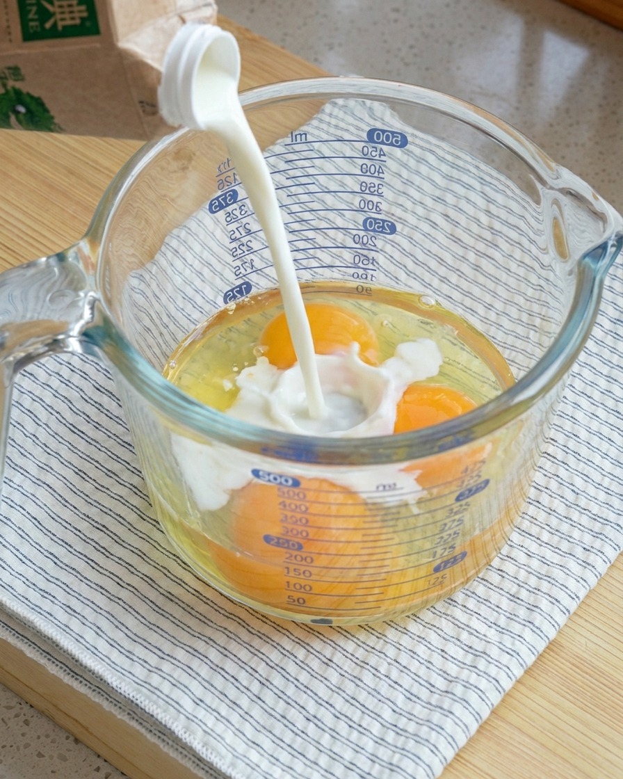 Milk being poured into a large glass measuring cup containing three cracked raw eggs.