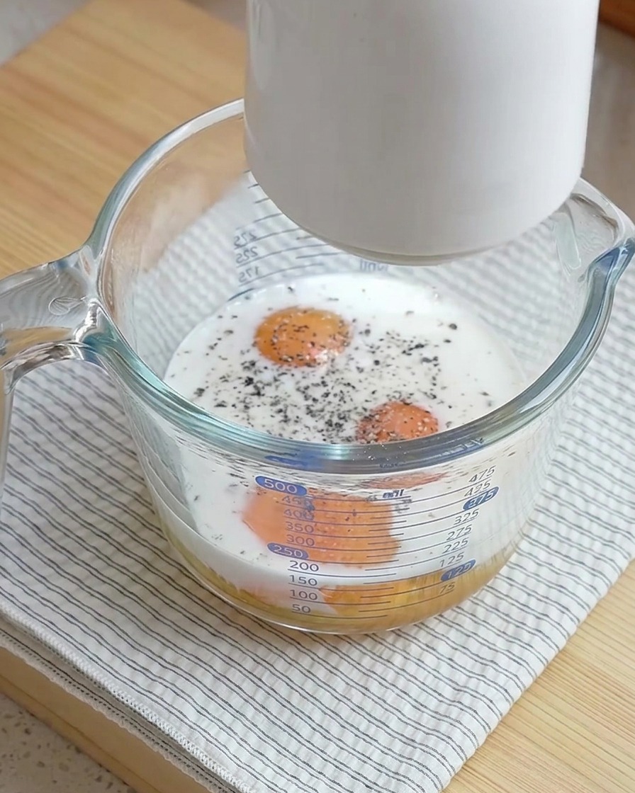 A white frothing tool held above a glass measuring cup filled with raw eggs, milk, and freshly ground black pepper.