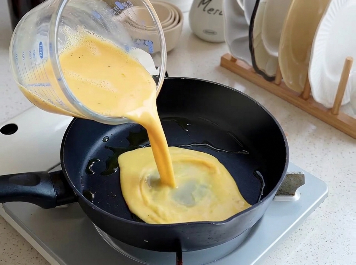 Beaten yellow egg mixture being poured from a glass measuring cup into a lightly oiled black frying pan.