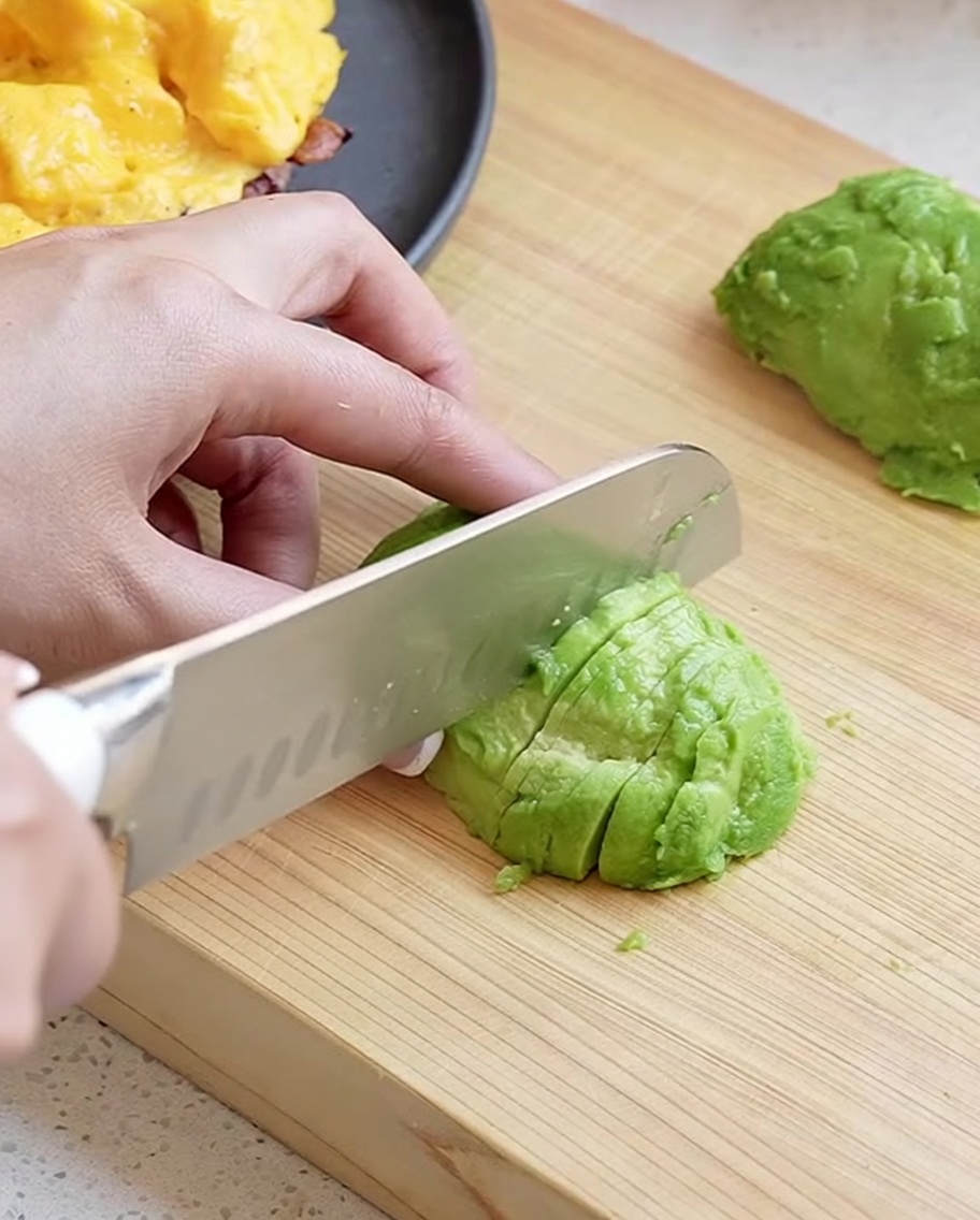 A hand using a chefs knife to slice a peeled avocado half into neat, thin pieces on a wooden board.