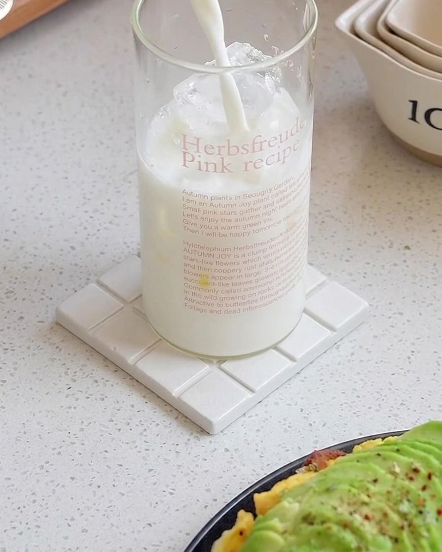 Milk being poured into a tall glass filled with ice cubes on a white coaster.