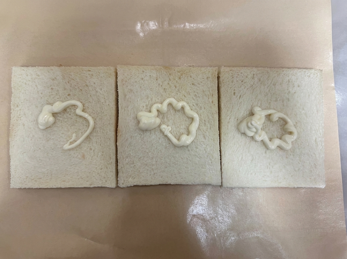 Three slices of white crustless bread laid out side-by-side on brown parchment paper, each topped with a squiggly line of white salad dressingcite: 1.