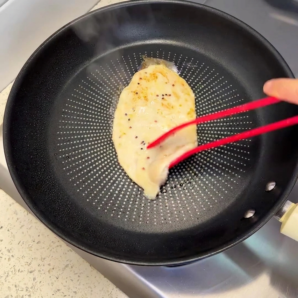 Red tongs turning over a partially cooked chicken patty in a frying pan, showing a browned crust.