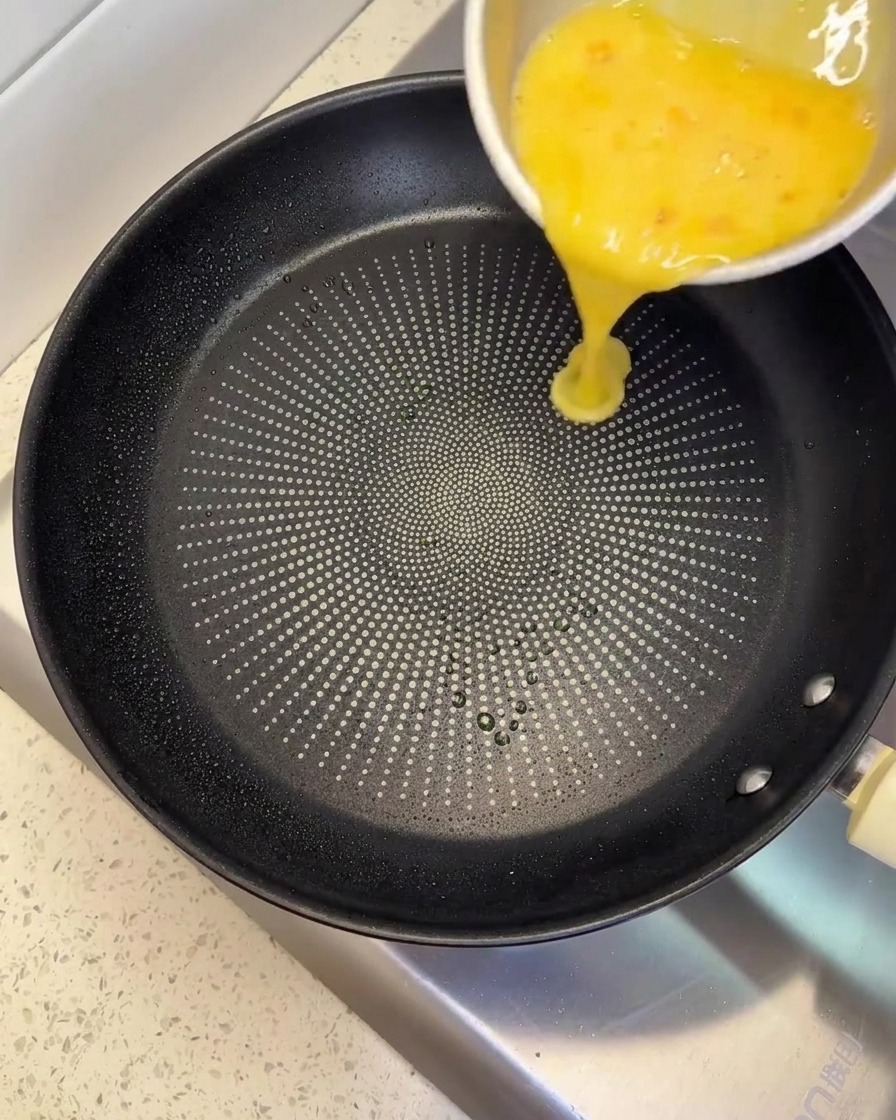 Pouring a yellow beaten egg mixture from a metal bowl into a black non-stick pan.