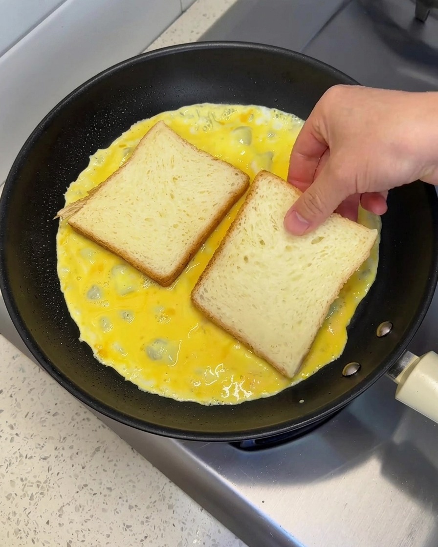 A hand placing two slices of white bread side-by-side onto a wet egg omelet cooking in a pan.