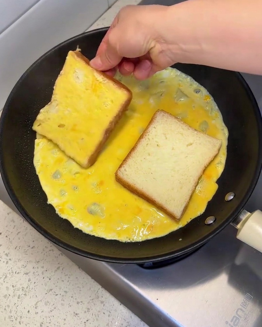 A hand placing and flipping a slice of bread in a hot pan filled with raw beaten egg to coat both sides.