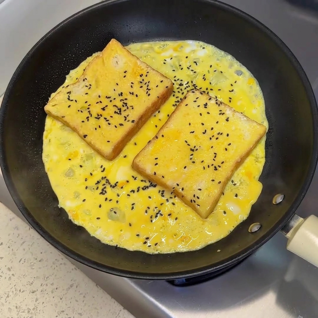 Black sesame seeds sprinkled over two slices of bread sitting in a partially cooked egg omelet in a frying pan.