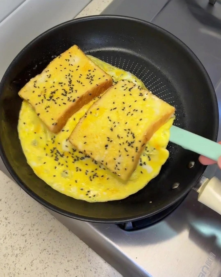 A silicone spatula lifting and flipping a large egg omelet containing two slices of bread in a pan.