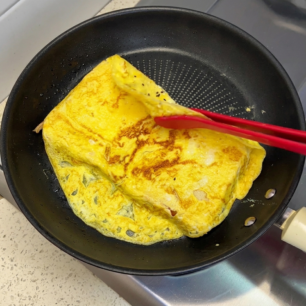 Red silicone tongs folding the overhanging edges of a cooked egg omelet onto two slices of bread in a pan.