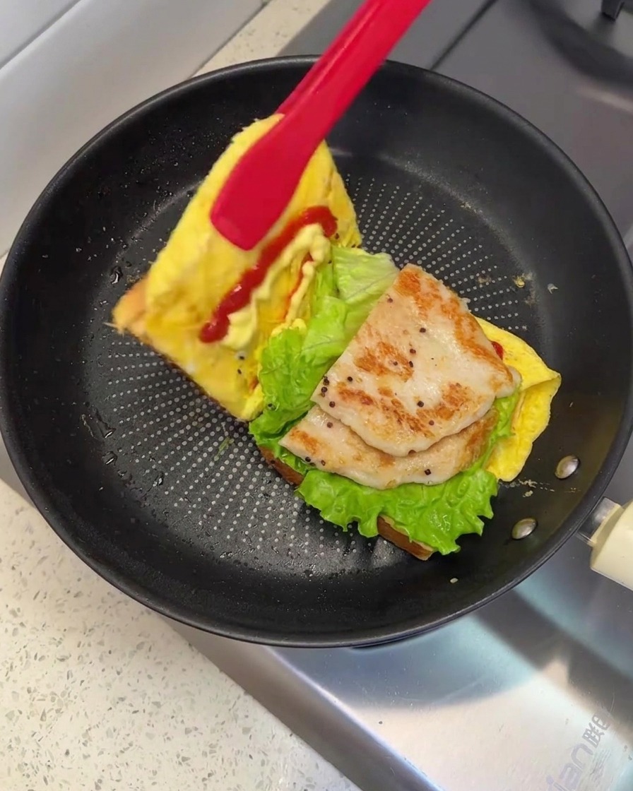 A pair of red tongs lifting and folding one side of the bread over to close the sandwich in the pan.