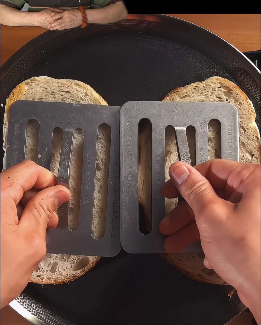 Slices of bread being pressed down and toasted in a black skillet until browned.
