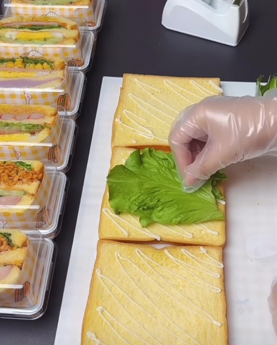A gloved hand carefully placing a fresh, green lettuce leaf on a slice of toast that has been drizzled with salad dressing.