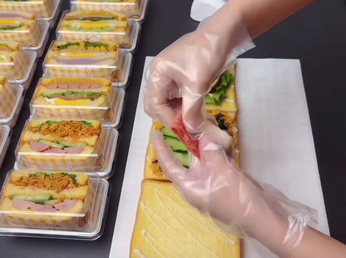 Gloved hands holding and arranging small sections of cooked bacon to place onto the sandwich build.