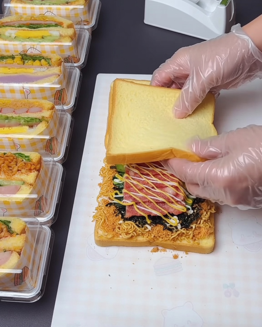 A pair of gloved hands lifting and stacking the top slice of toast onto the loaded layers below.