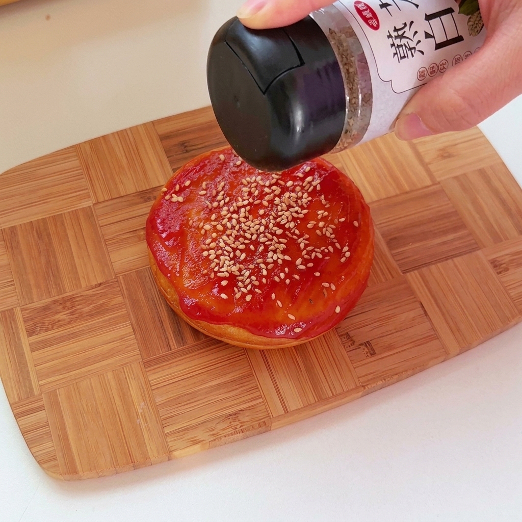 A close-up of ketchup being applied to the top of a savory round bun with sesame seeds.