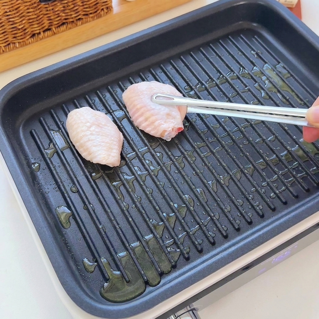 Raw chicken wings being placed onto a ribbed grill pan with oil using tongs.