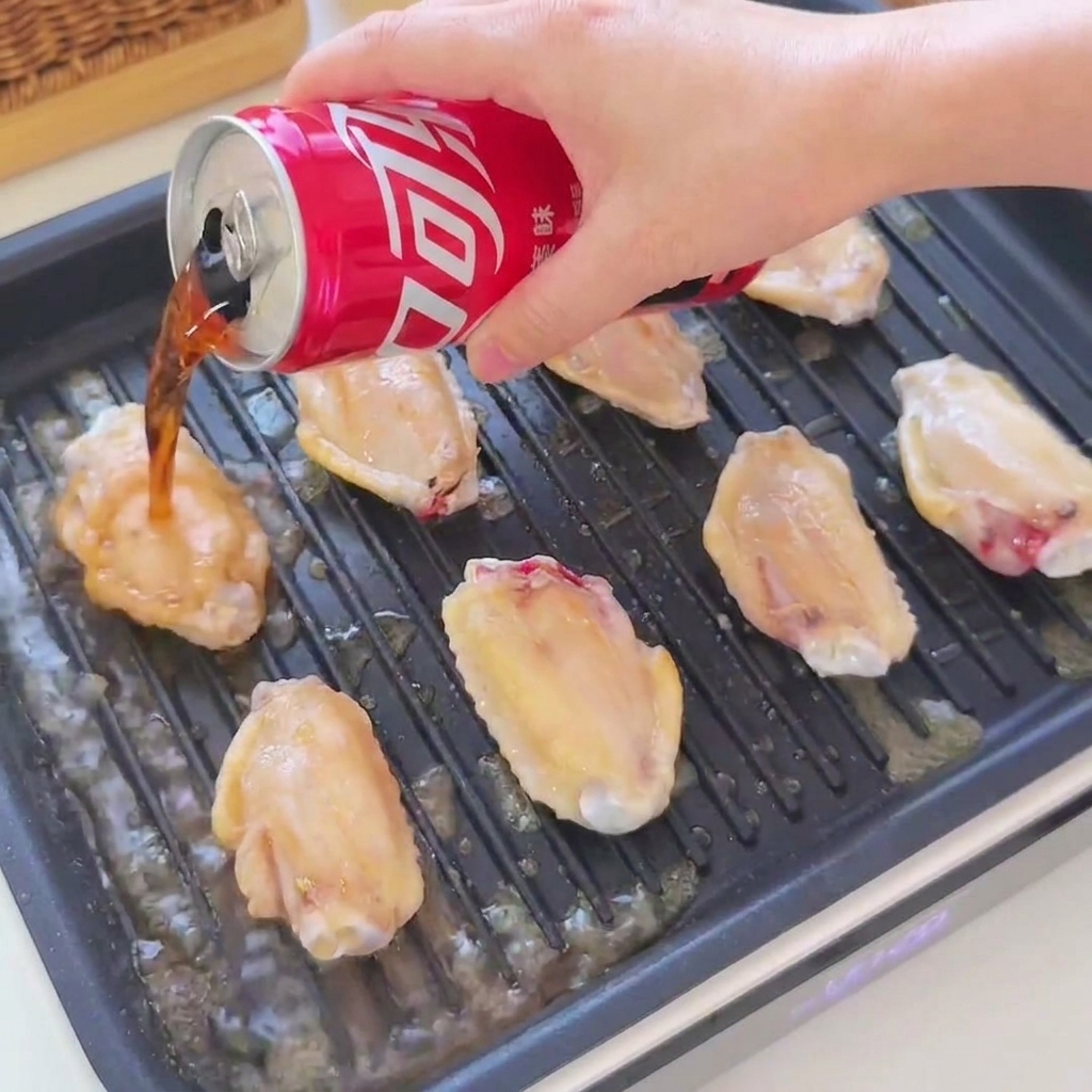 Cola and soy sauce being poured from a red can over golden chicken wings sizzling in a black grill pan.