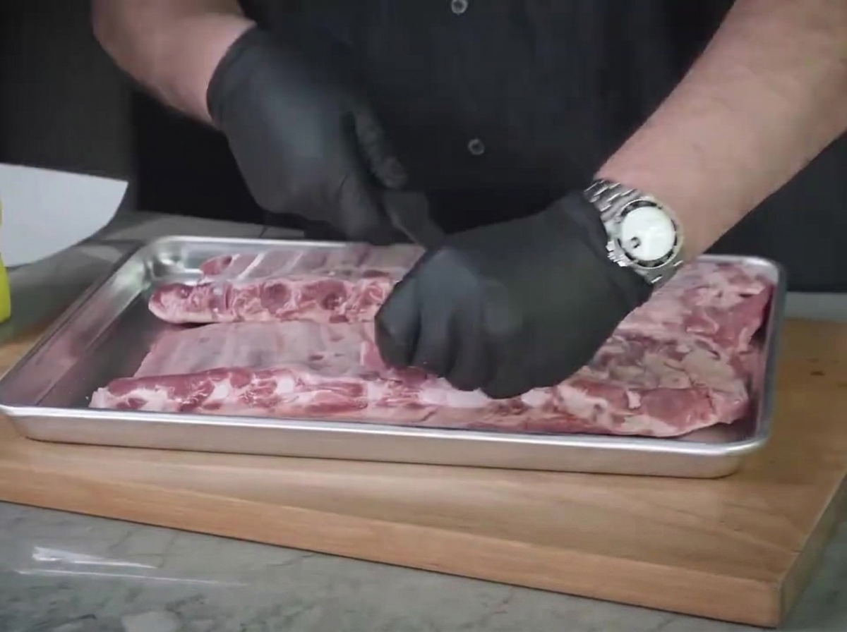 A close-up view of gloved hands cleaning and trimming excess fat off raw St. Louis ribs on a steel tray.