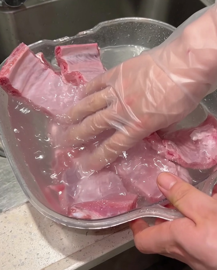 Gloved hands scrubbing and washing raw pork ribs inside a water-filled container.