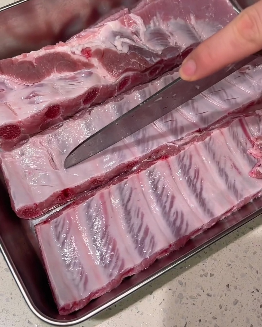 A sharp knife slicing shallow cuts across the fascia of raw pork ribs on a metal tray.