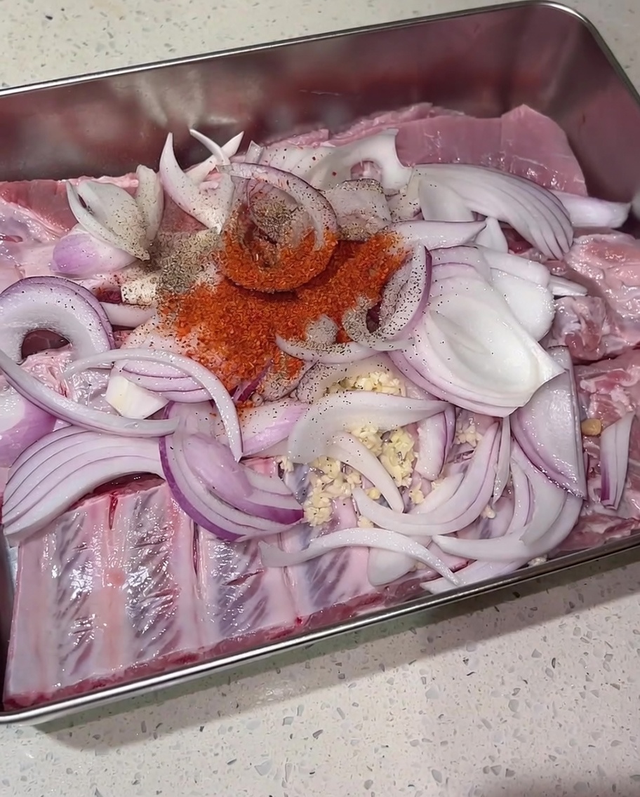 A pile of red chili powder, dark black pepper, and brown sugar sitting on top of raw pork ribs and sliced red onions in a metal tray.
