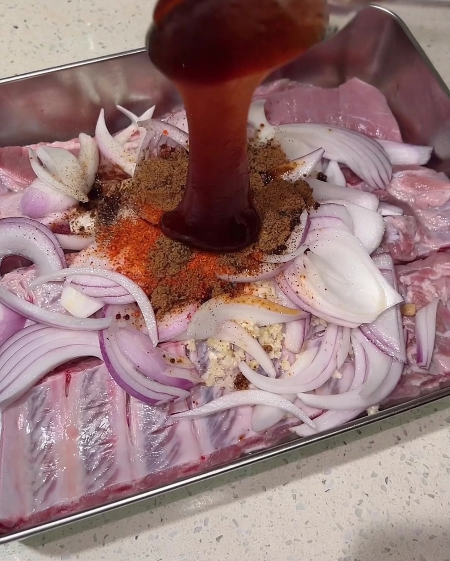 Thick, dark red char siu sauce being poured from above onto the mound of dry spices, raw pork ribs, and onions.