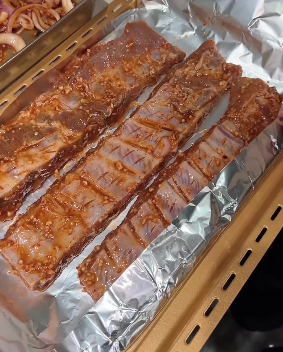 Three strips of marinated pork ribs laid out on aluminum foil over a gold baking tray, ready to be wrapped.