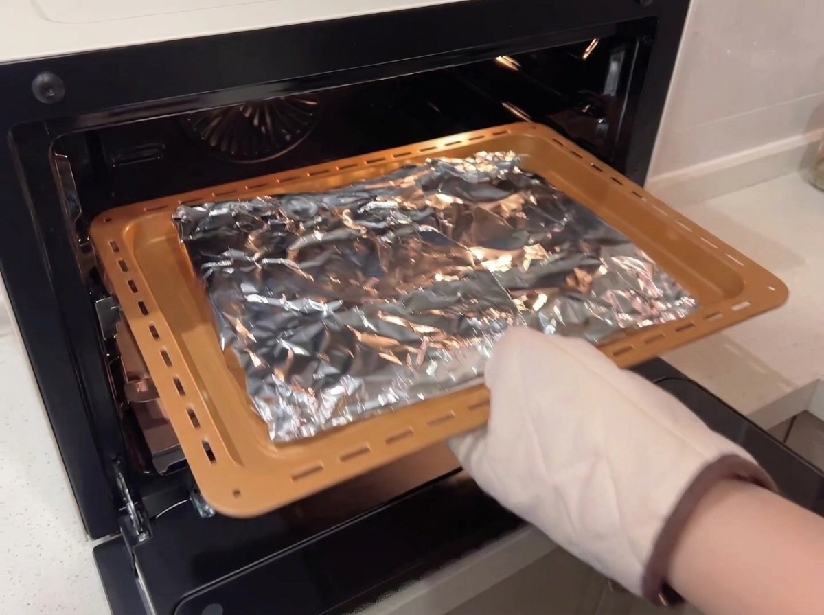 A person wearing a protective oven mitt placing a baking tray with foil-wrapped ribs into a convection oven.