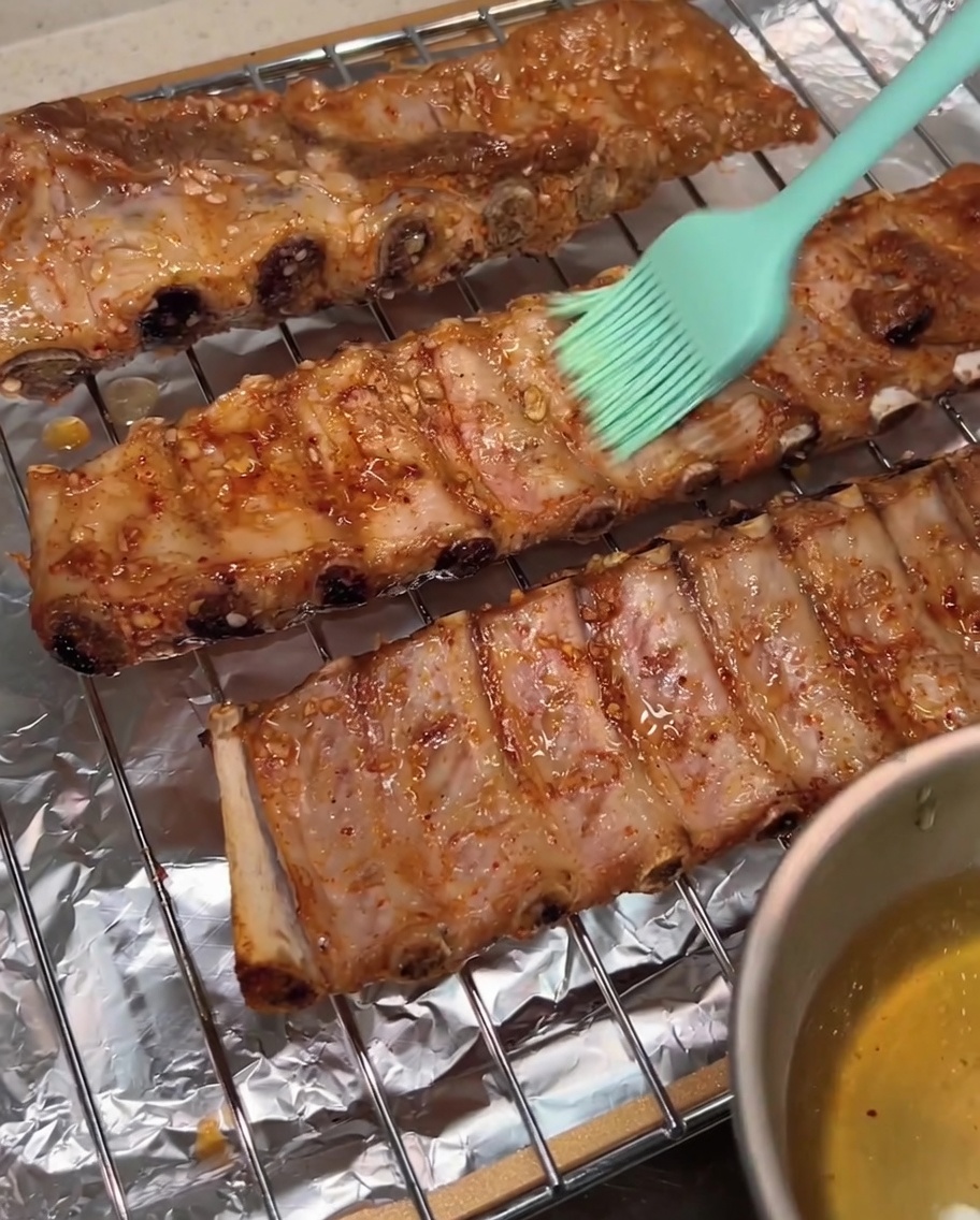 A green silicone brush applying a glossy coating of honey water onto the surface of the roasted pork ribs on a wire rack.