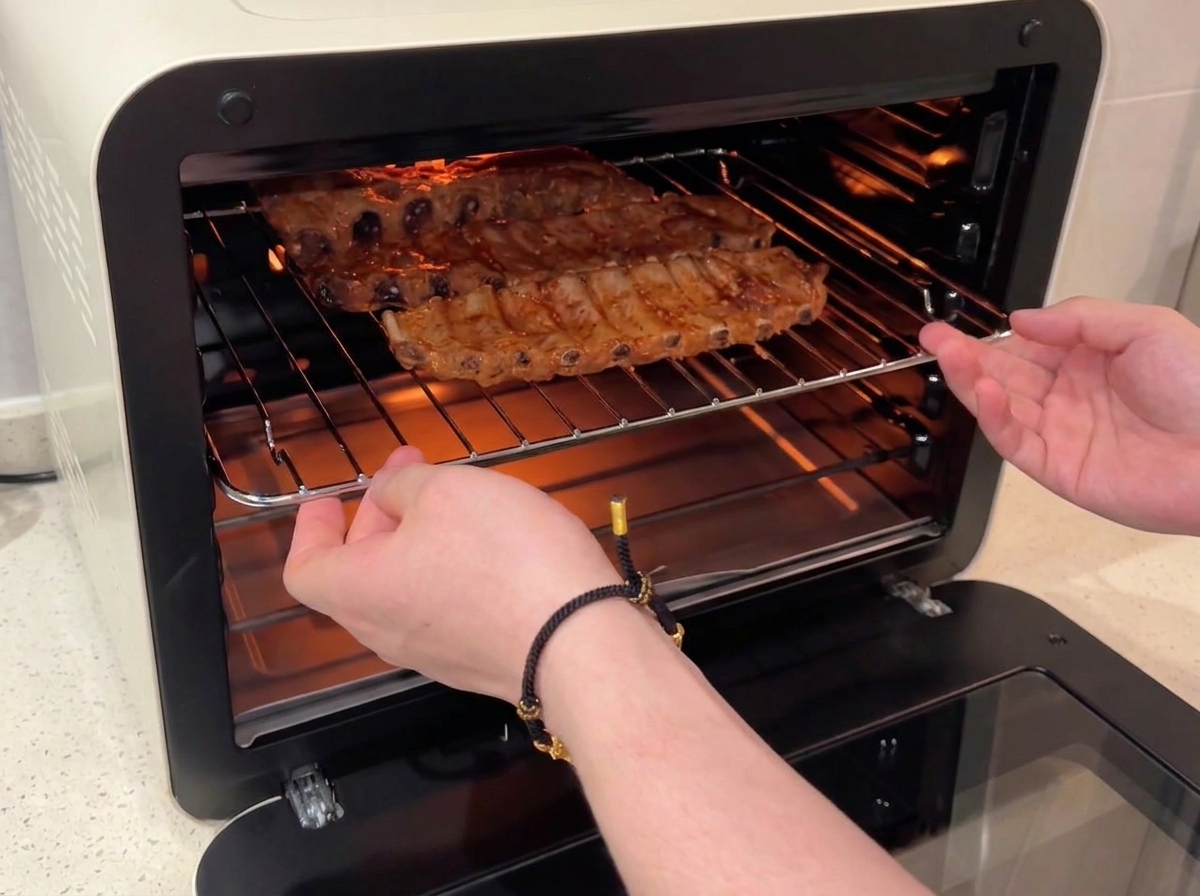 Hands sliding a wire rack holding honey-glazed pork ribs back into a lit convection oven.