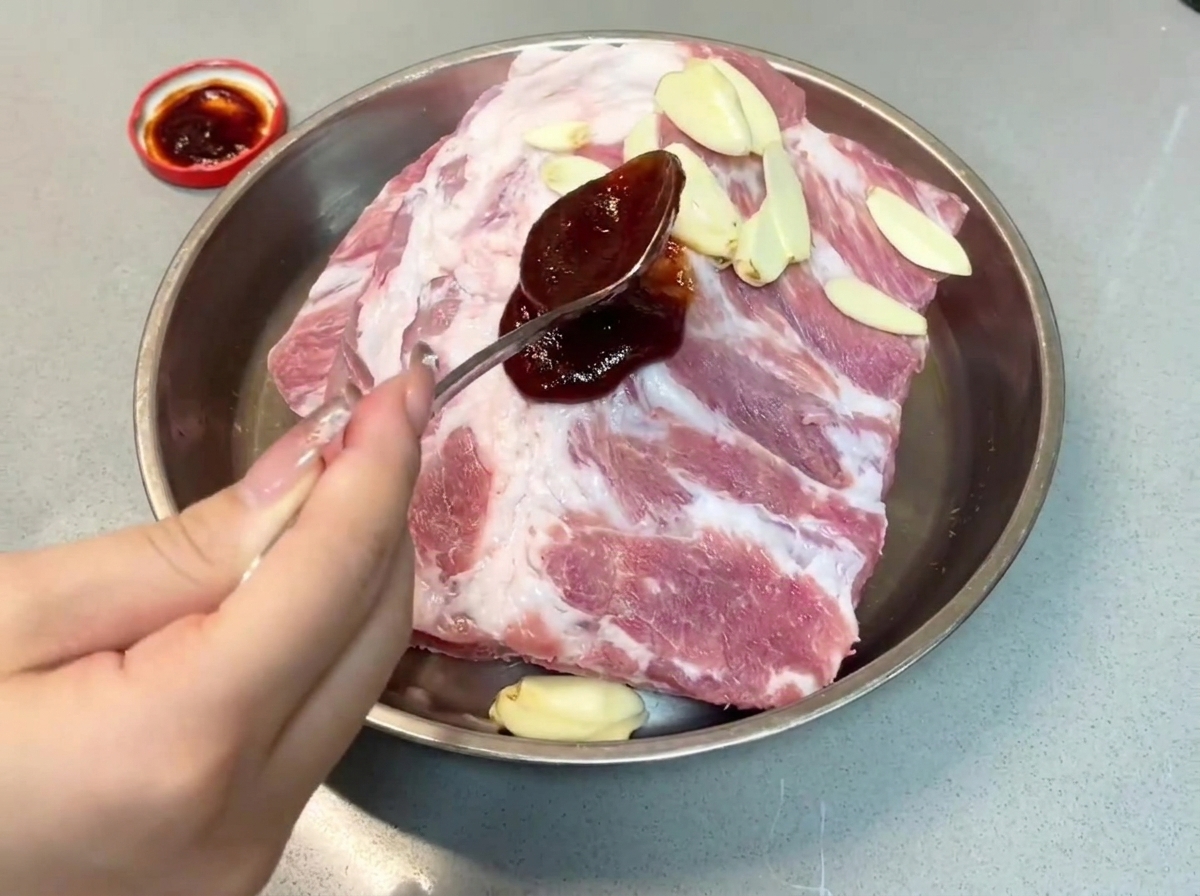 A spoonful of dark char siu sauce being poured onto pork ribs in a metal tray.