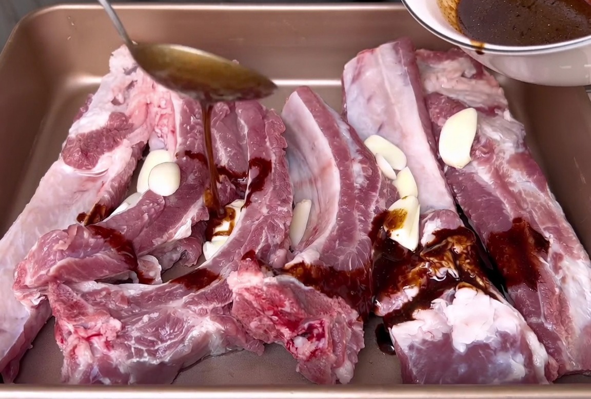 Pouring a dark soy-based marinade from a white bowl over raw pork ribs and garlic slices in a baking pan.