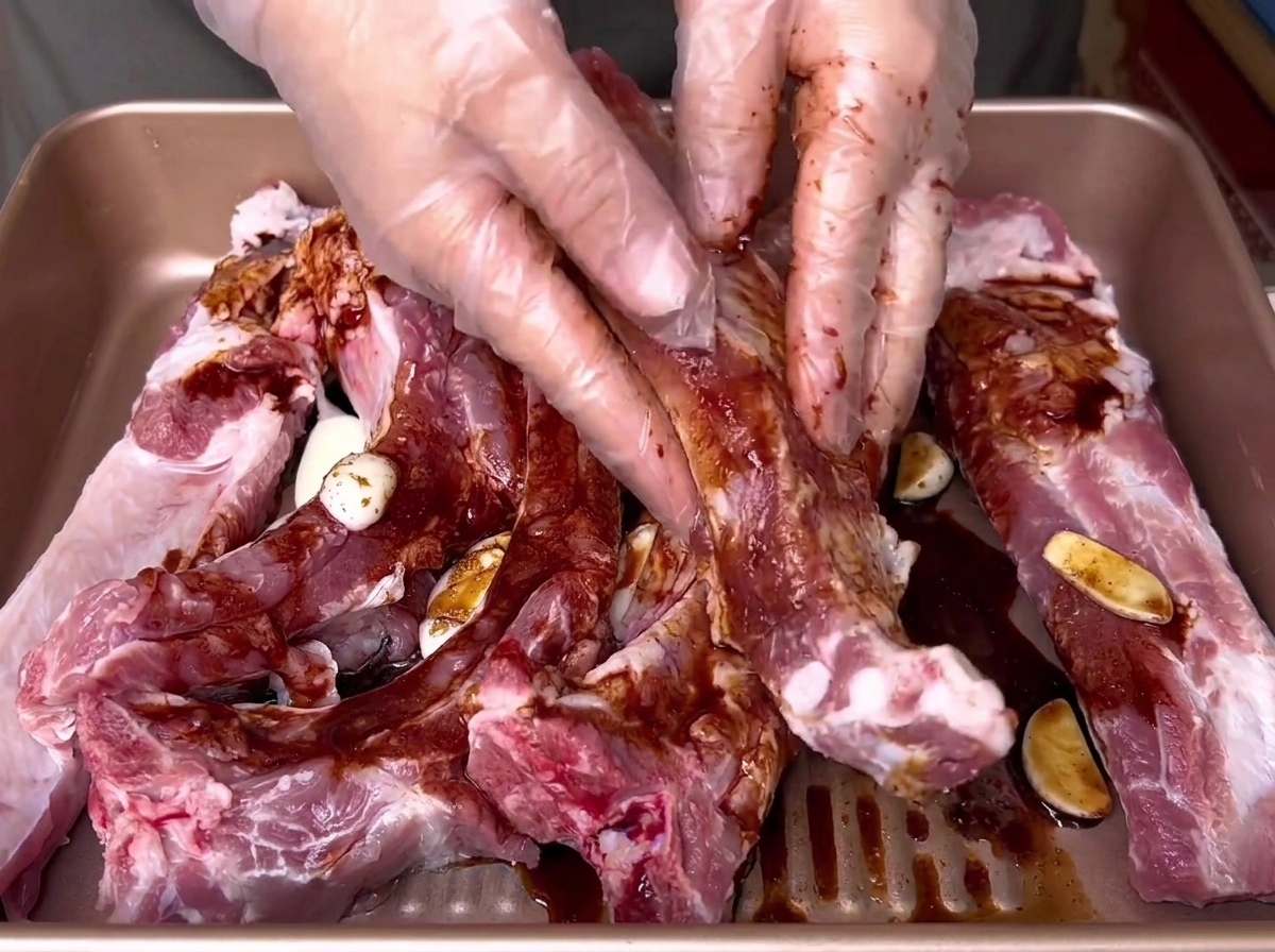 Gloved hands thoroughly rubbing a dark liquid marinade into raw pork ribs inside a golden tray.