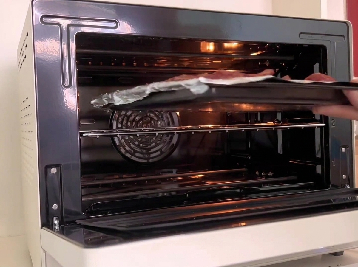 A baking tray holding foil-wrapped ribs being slid into the middle rack of a lit white oven.
